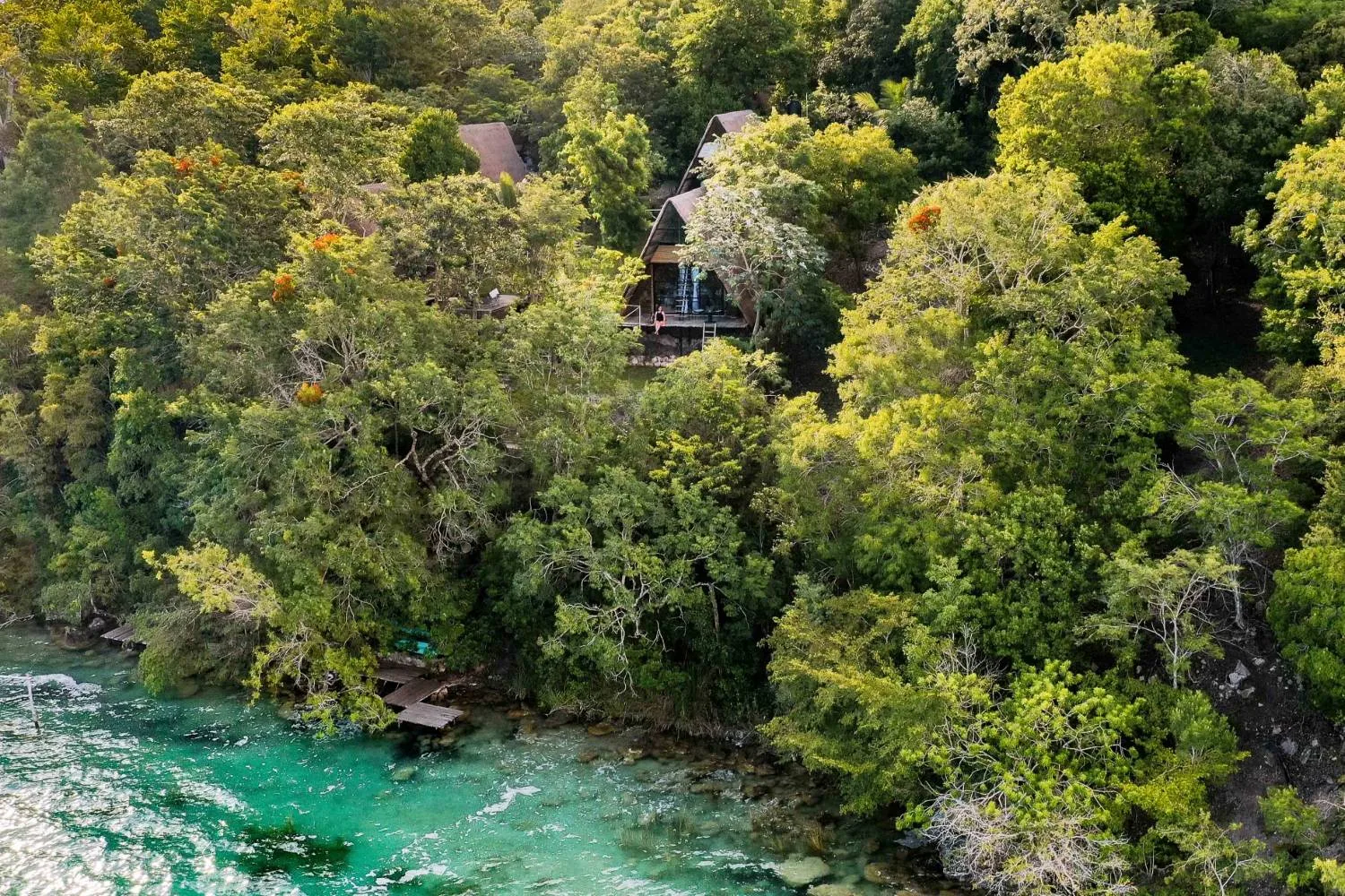 Aerial view of A-frame cabins among the forest above the shore of Bacalar lagoon, with a wooden dock and turquoise water