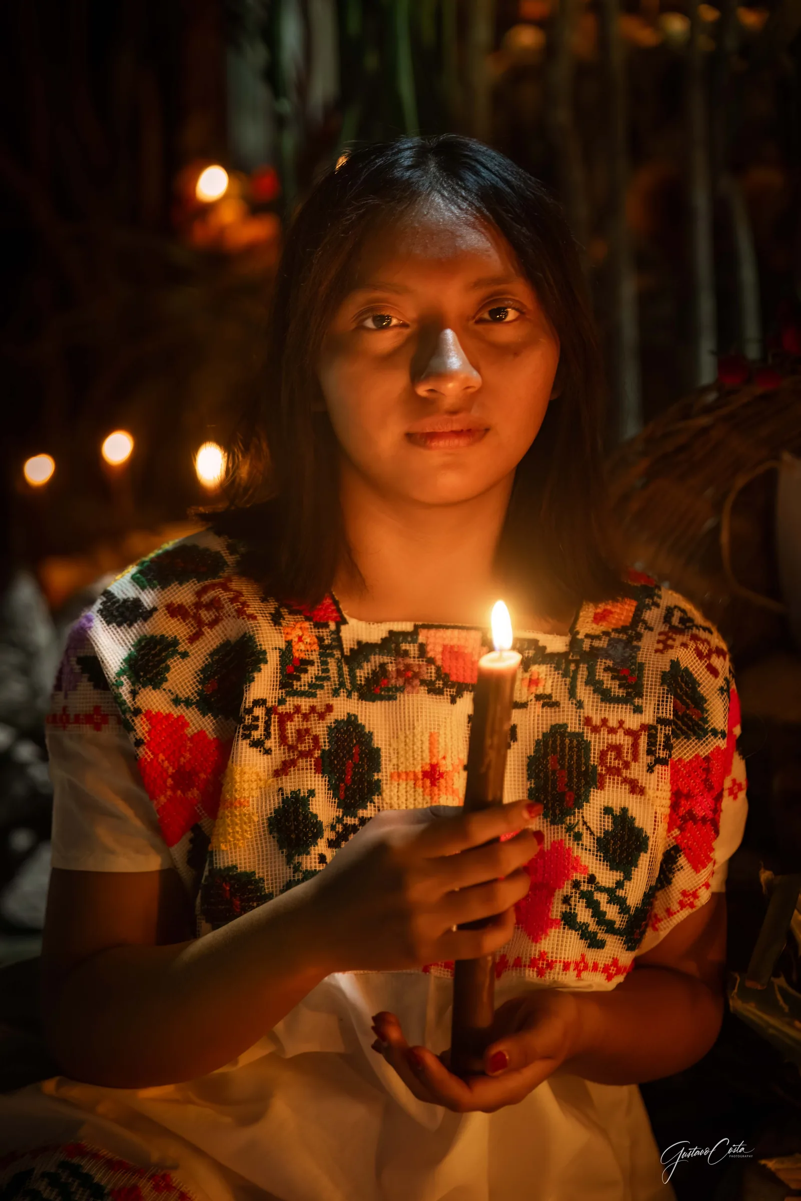 Young woman in an embroidered huipil holding a lit candle before a Hanal Pixán altar