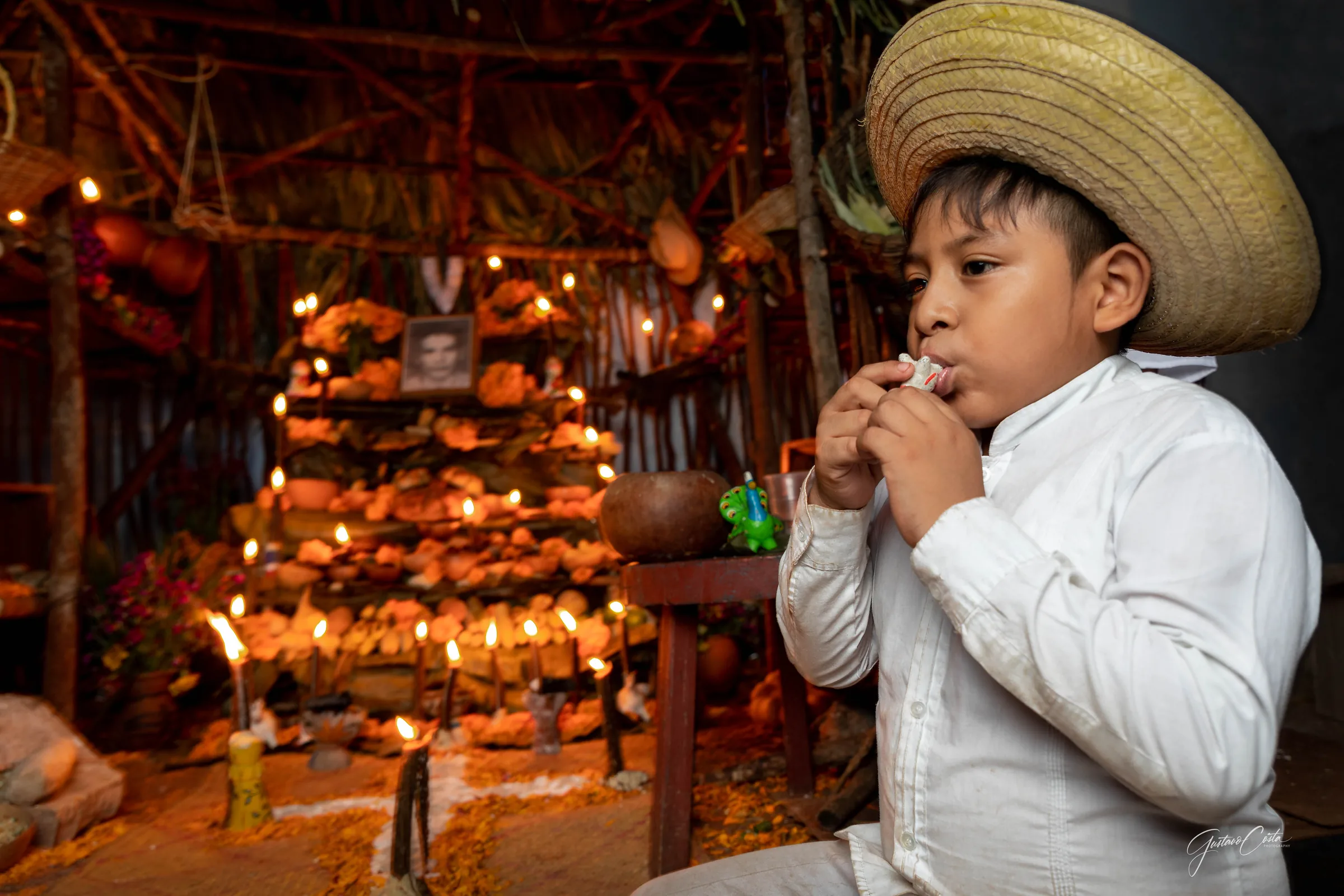Boy in a palm hat playing a clay whistle beside a candlelit Hanal Pixán altar